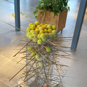 Hoge cilindervormige draadmand gevuld met tennisballen, waarin houten stokken horizontaal zijn gestoken om de ballen op hun plek te houden.
