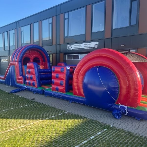 Opblaasbare stormbaan van 17,5 meter in rood-blauwe kleuren, met diverse hindernissen zoals klimdelen, kruiptunnels en obstakels, opgesteld op een witte achtergrond.