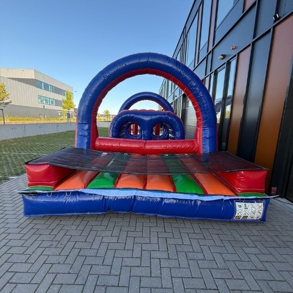 Een rood-blauwe stormbaan van 10 meter lang met diverse hindernissen, waaronder een tunnel, obstakels en een klim-/springgedeelte, geplaatst op een kleurrijk opblaasbaar platform.